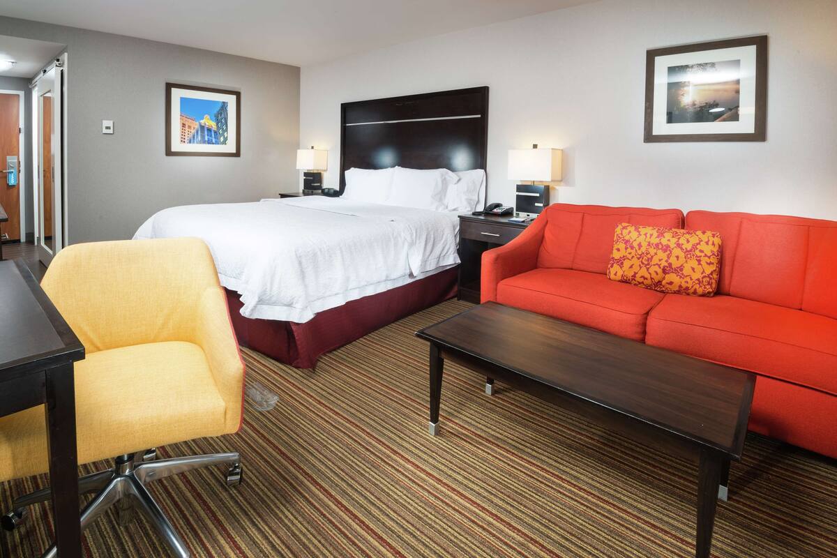 A hotel room with a bed, a red sofa, a yellow chair, a desk, and a small table.