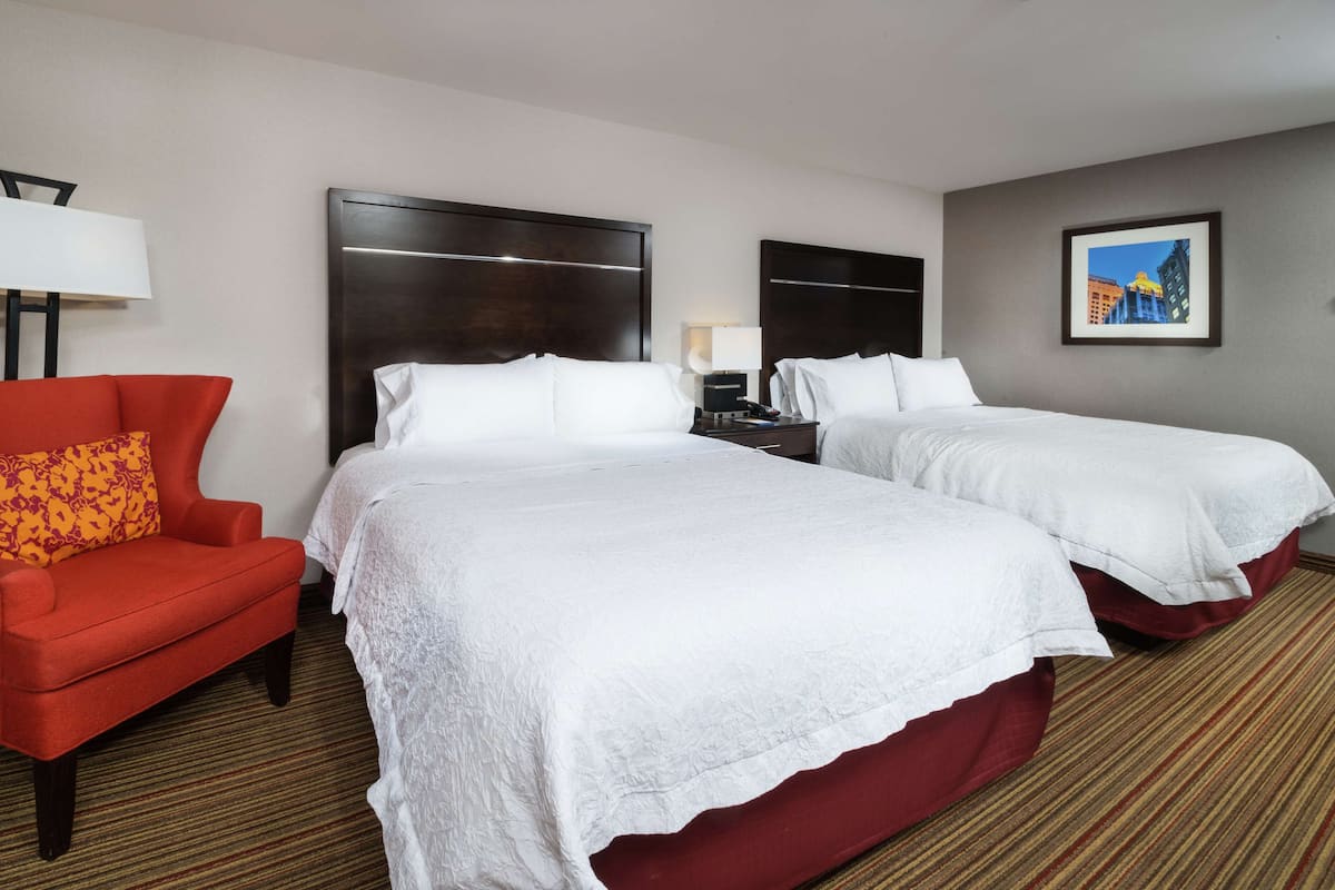 A hotel room with two beds, a red armchair, a nightstand, and a framed picture on the wall.