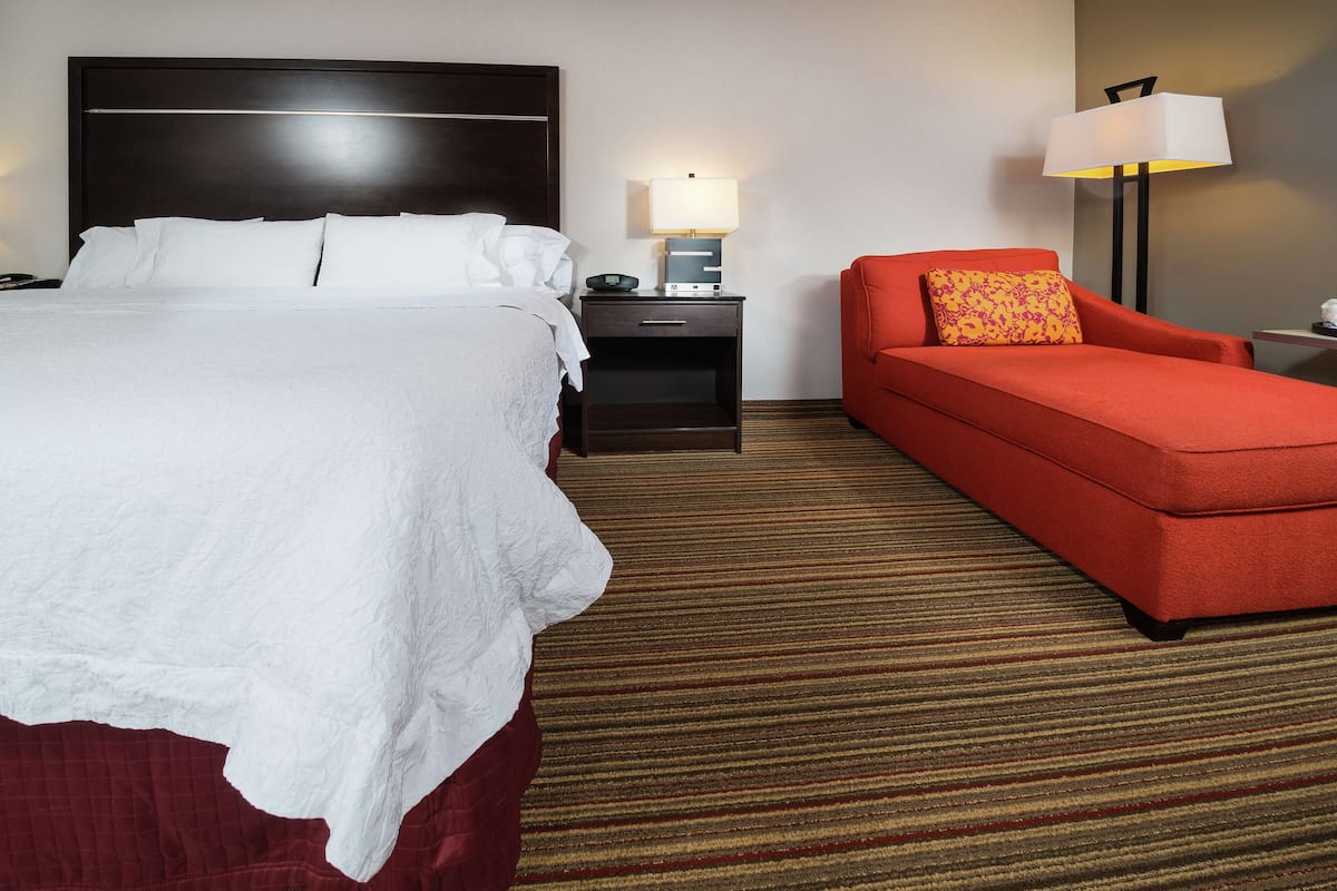 A hotel room with a large bed, a red armchair, a nightstand with a lamp, and a carpeted floor.