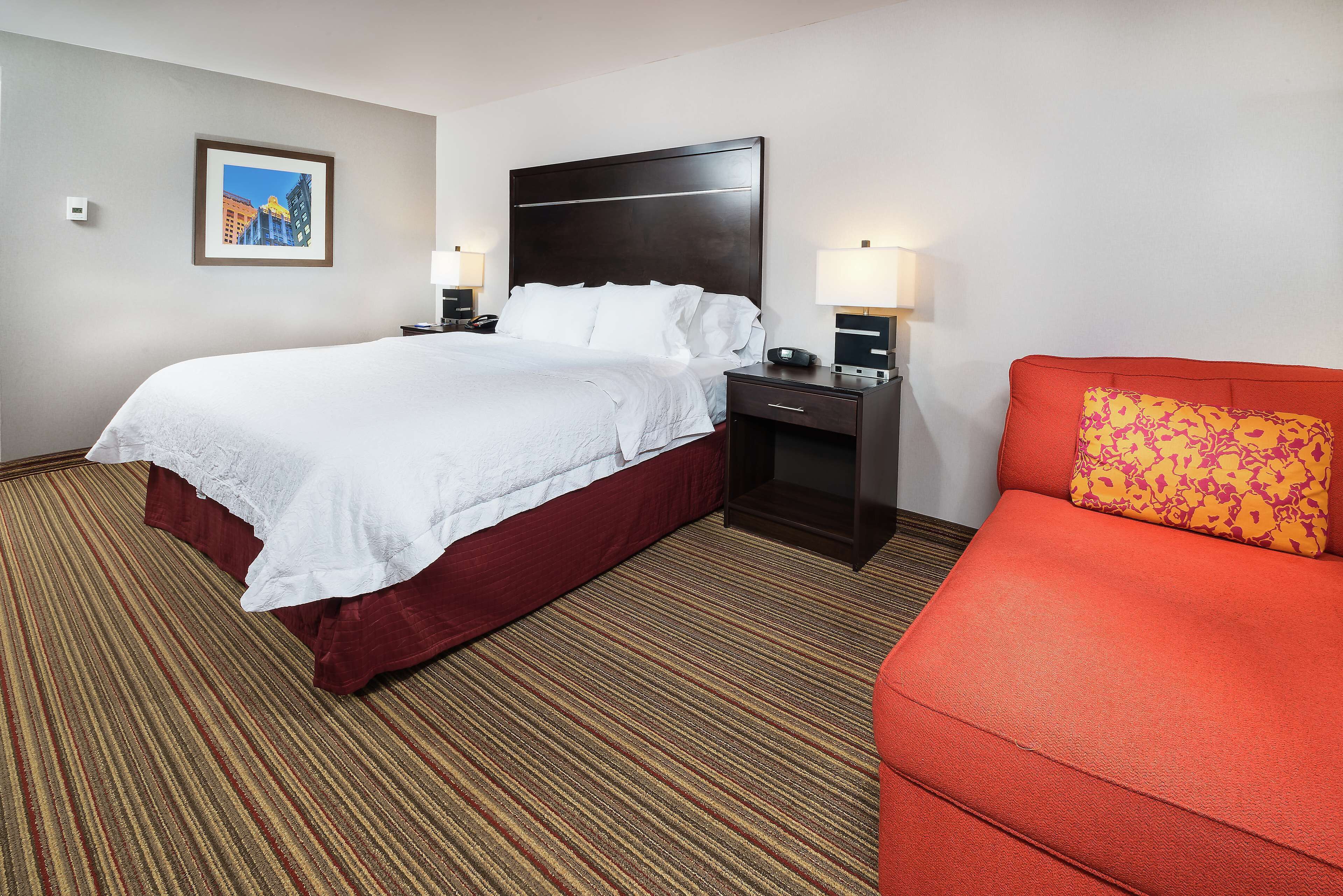 A hotel room with a large bed, a red sofa, a nightstand with a lamp, and a framed picture on the wall.