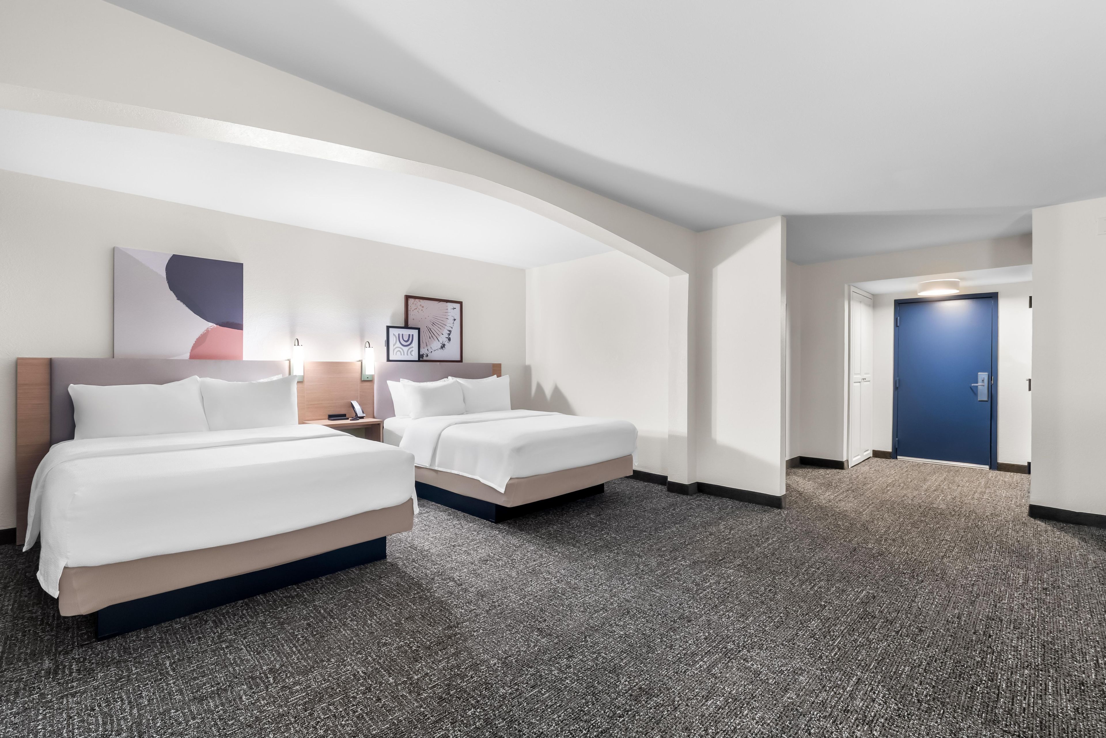 A hotel room with two beds, a desk, a chair, a phone, and a door.