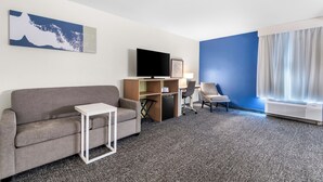 A hotel room with a grey sofa, a small white side table, a desk with a computer and a chair, a flat-screen TV, and a large window with curtains.