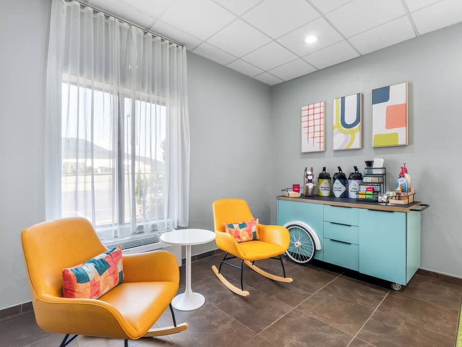 A modern room with two orange chairs, a small white table, a teal cabinet with coffee and water dispensers, and abstract art on the wall.