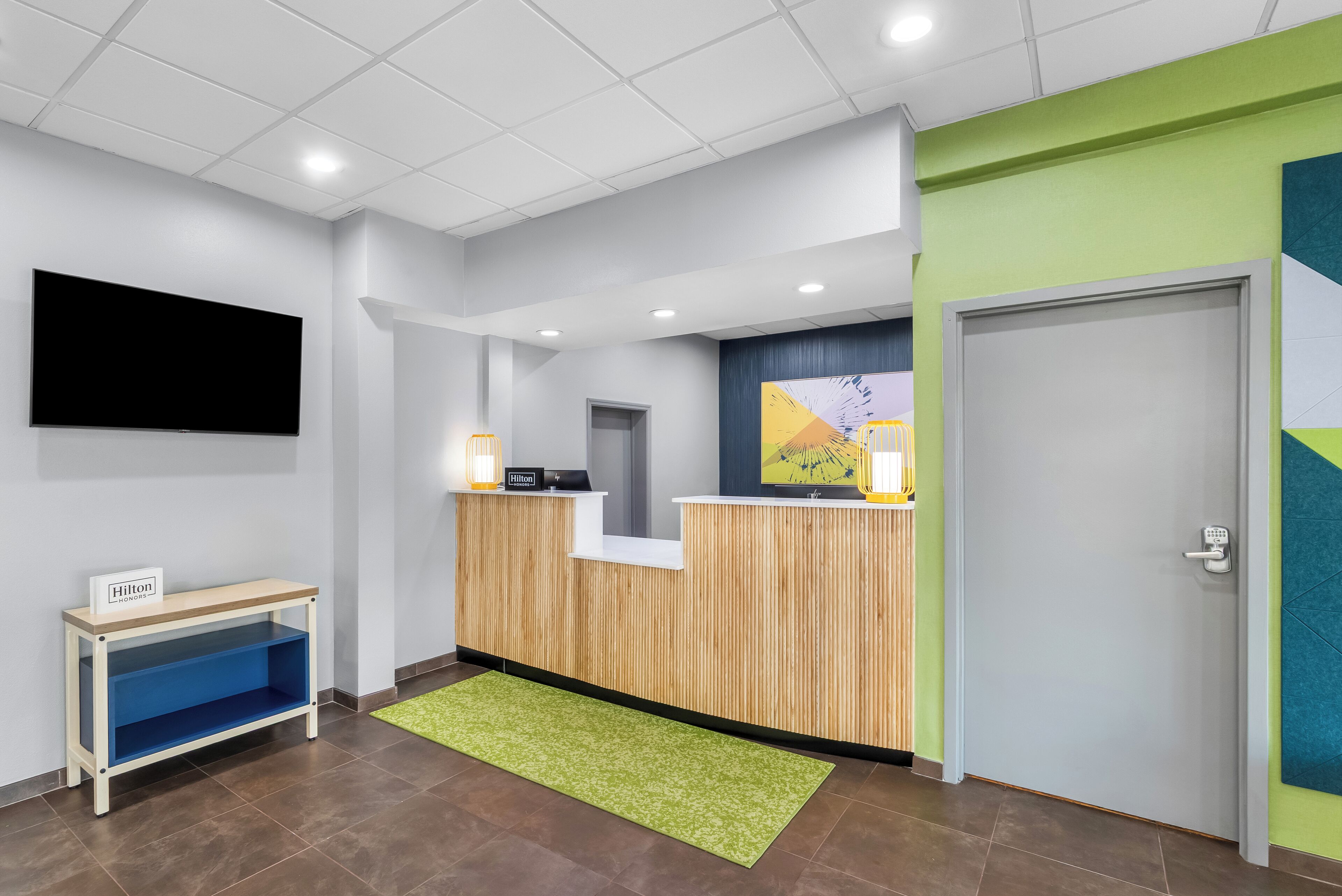 A hotel reception area with a flat-screen TV, a wooden reception desk, a console phone, a digital display, a small blue seating area, and a green rug.