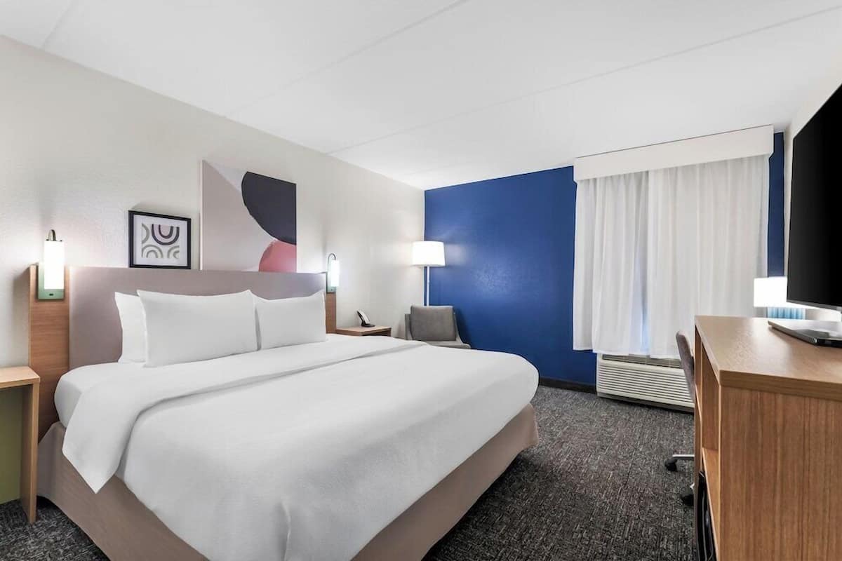 A hotel room with a bed, a desk, a chair, a lamp, a picture frame, and a window with curtains.