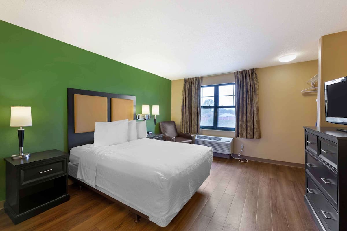 A hotel room with a large bed, a nightstand with a lamp, a chair, a dresser, and a television.
