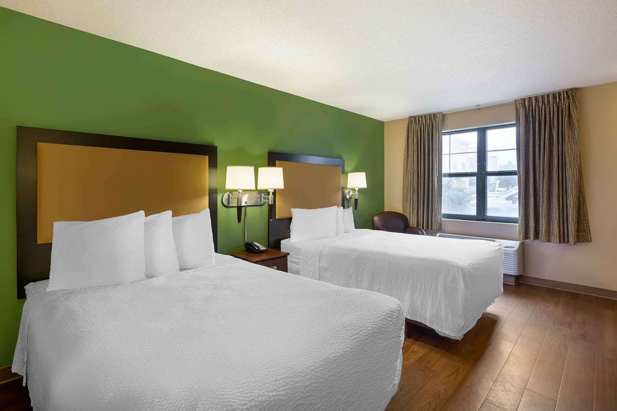 A hotel room with two beds, green walls, a large window with curtains, and a nightstand with a lamp.