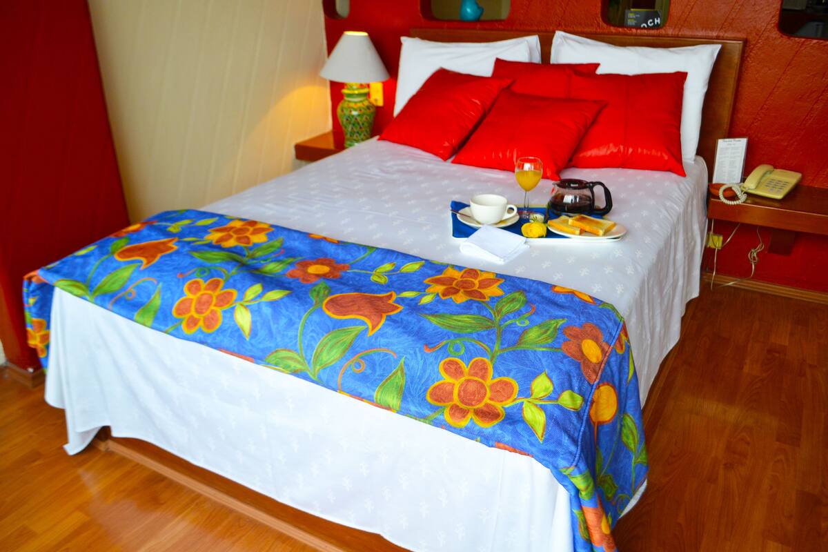 Un lit avec une couvre-lit à motifs floraux, des oreillers rouges, une table de chevet avec une lampe et un téléphone.
