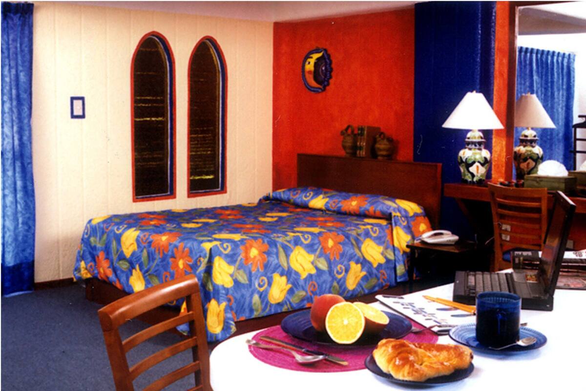 Une chambre d’hôtel avec un lit, un bureau et une chaise. Il y a une fenêtre avec des rideaux, une lampe et une horloge au mur.