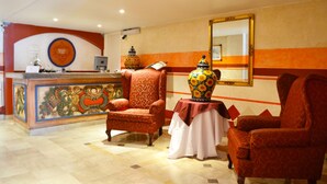 Un espace d’accueil avec un comptoir décoratif, deux fauteuils à motifs, une table avec un vase et un grand miroir au mur.