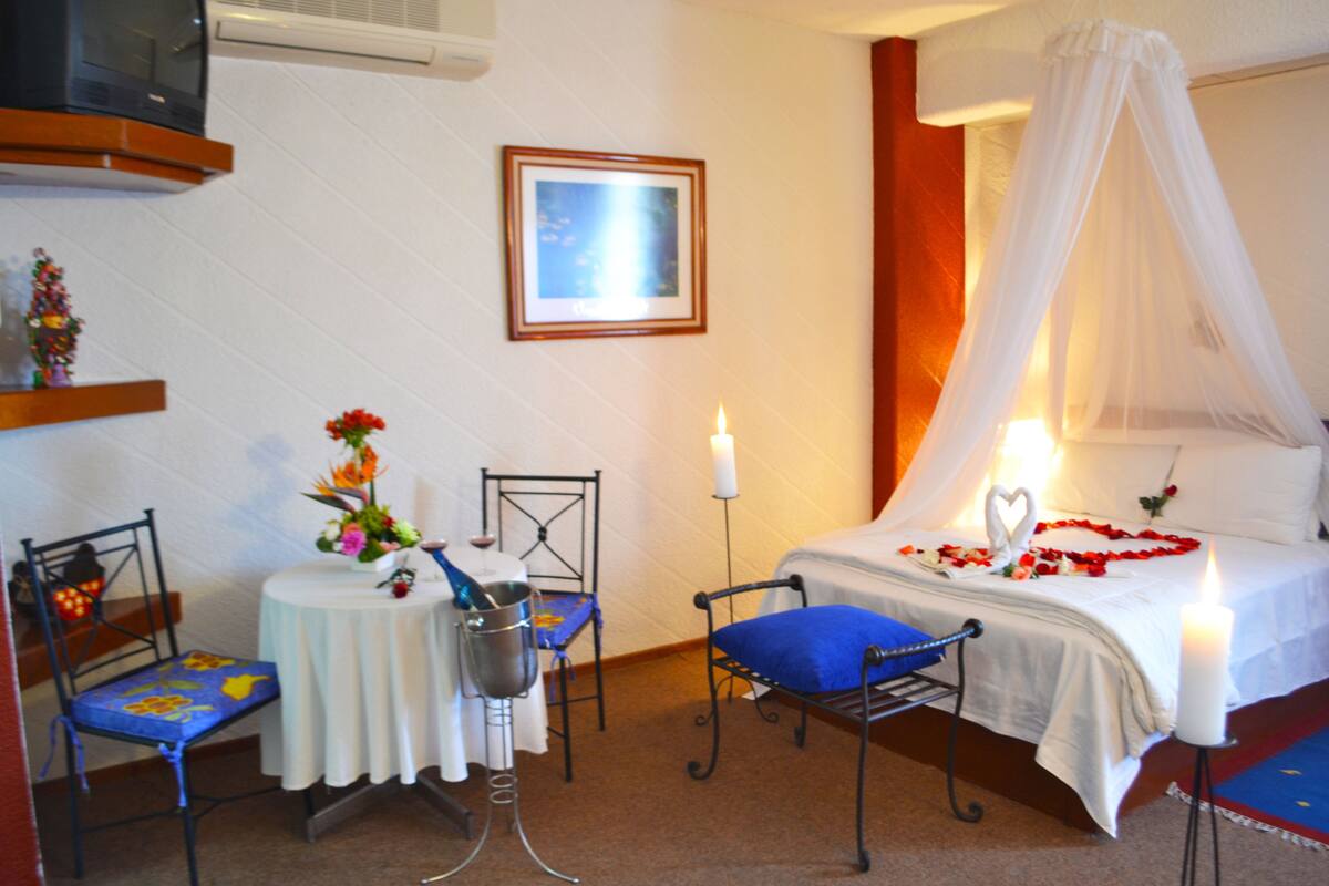 Une chambre à coucher avec un lit, une table de chevet, une chaise, une petite table ornée d’un vase de fleurs et un tableau accroché au mur.