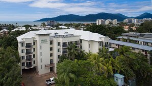 A multi-story building labeled "Tropic Towers" with balconies, surrounded by lush greenery and a view of the sea and mountains in the background. - Tropic Towers Apartments (Cairns North)
