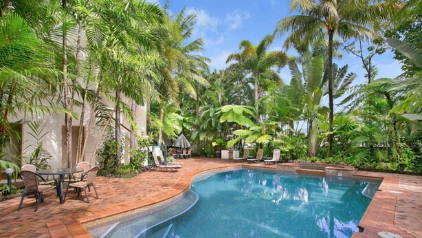 A swimming pool with a curved edge, surrounded by palm trees and a brick patio. - Tropic Towers Apartments (Cairns North)