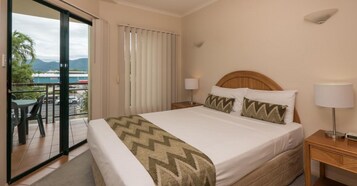 A hotel room with a bed, two bedside lamps, a view of a balcony with outdoor furniture, and a window with blinds.