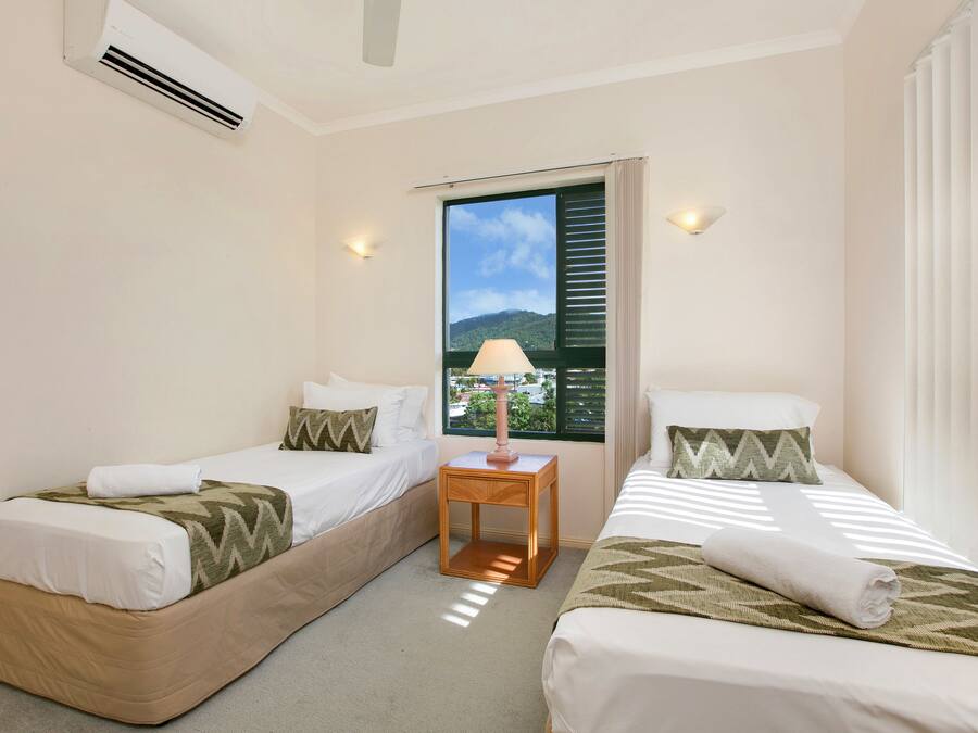 A hotel room with two single beds, a small wooden nightstand with a lamp, and a window with a view of a mountain and a cityscape.