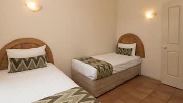 Two single beds with wooden headboards, white bedding, and patterned throw pillows in a room with tiled flooring and a closed door.