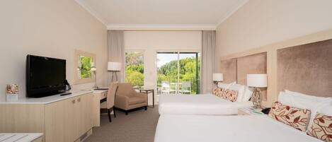 A hotel room with a TV, desk, two beds, and a balcony with a view of greenery.