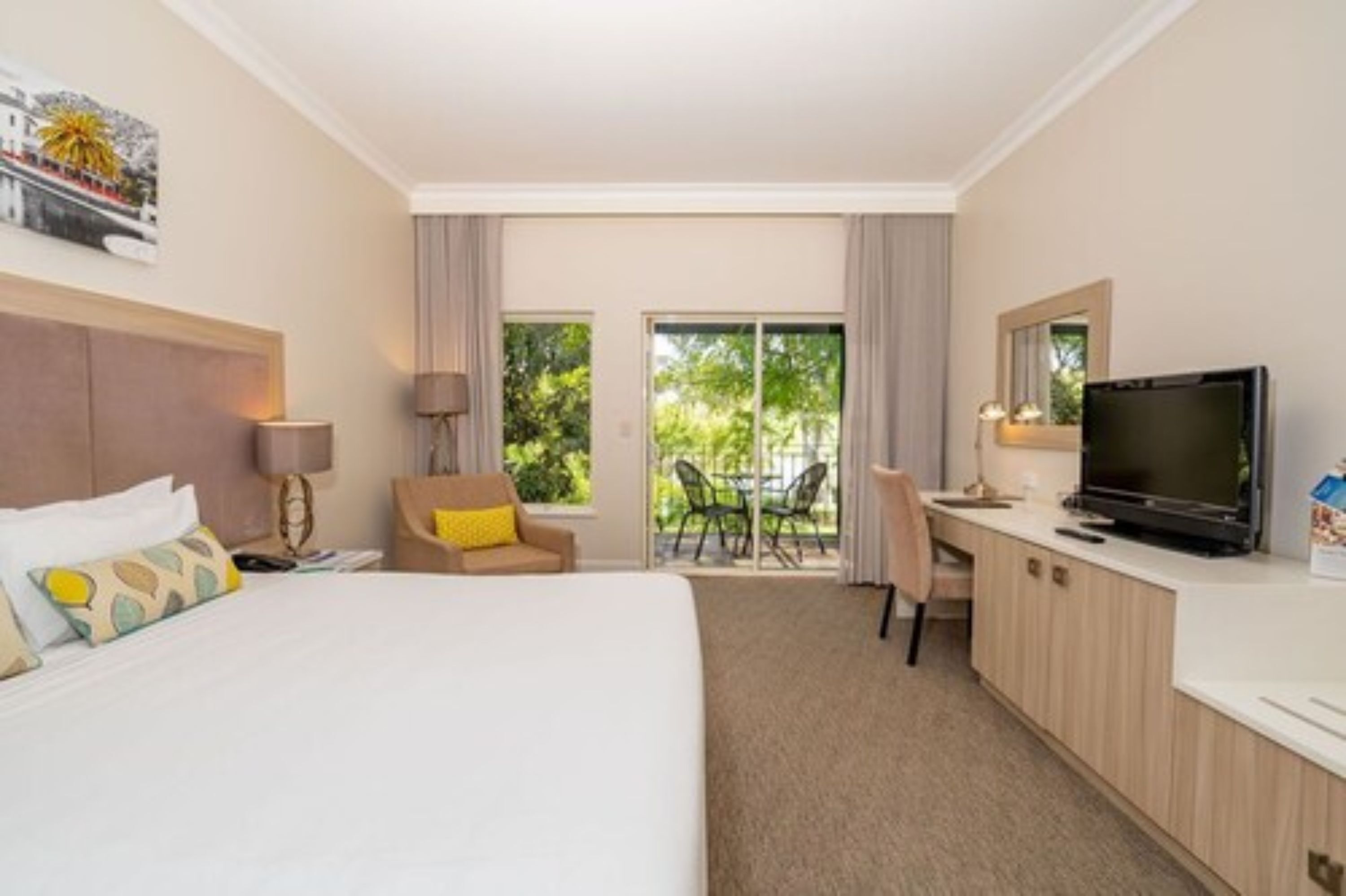 A hotel room with a bed, a desk, a television, and a balcony with a view of greenery.
