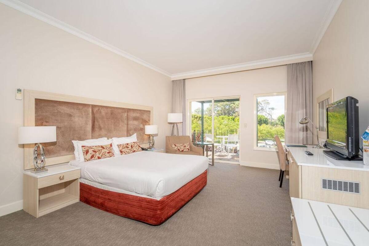 A hotel room with a large bed, two bedside tables with lamps, a desk with a TV, and a view of greenery through the windows.