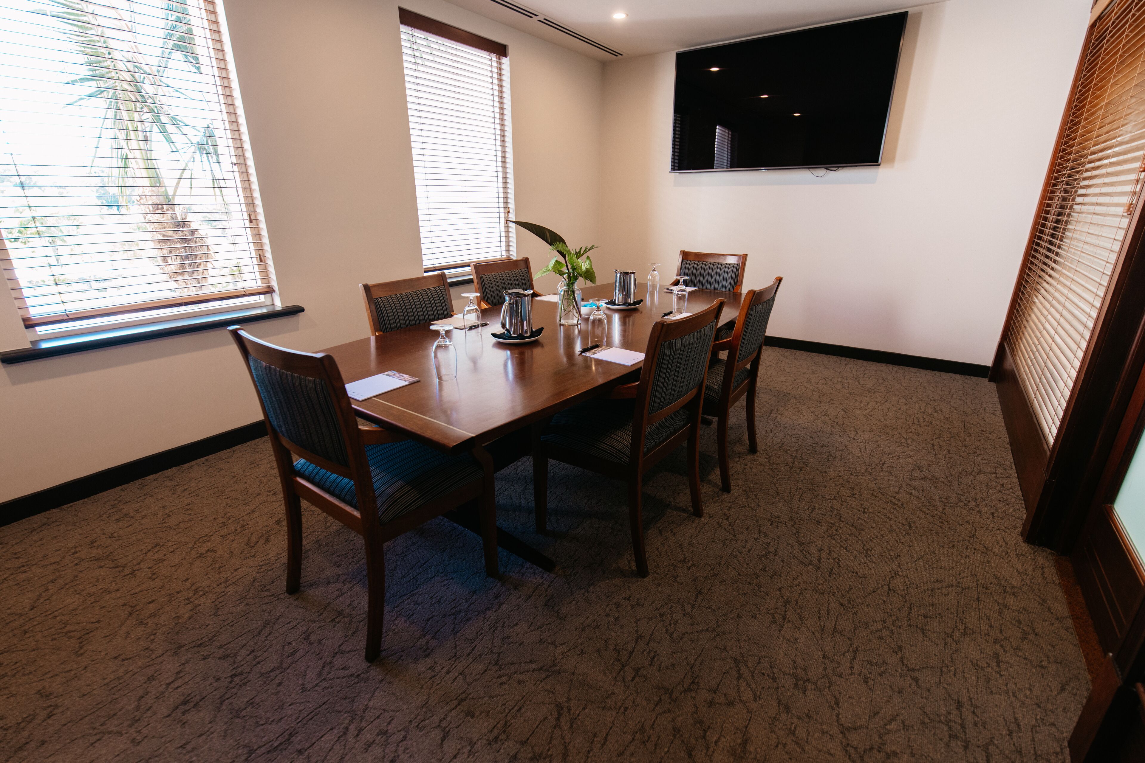 A conference room with a wooden table, six chairs, a large window with blinds, a mounted TV, and a carpeted floor.