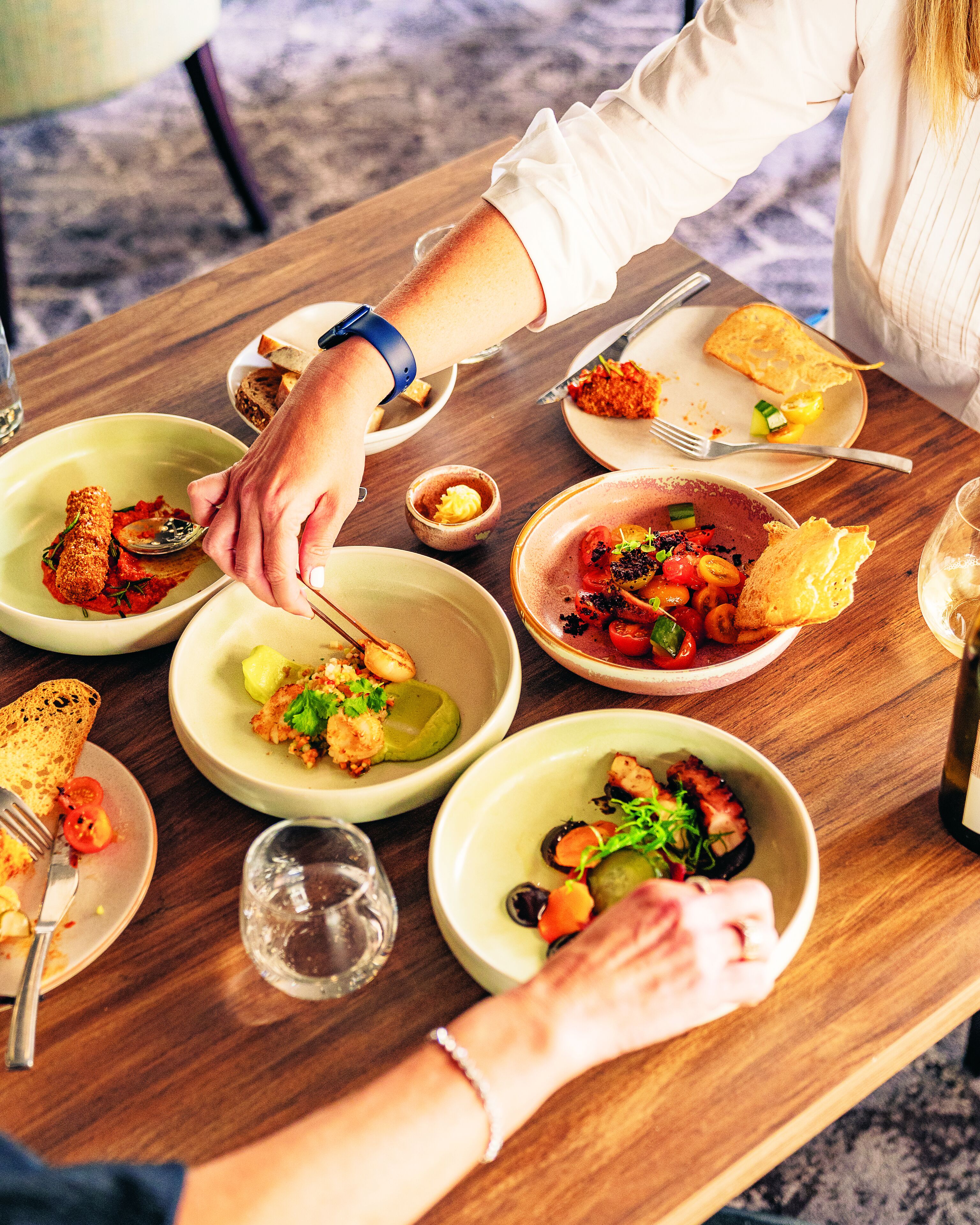 Une table à manger avec différents plats, couverts et un verre d’eau.