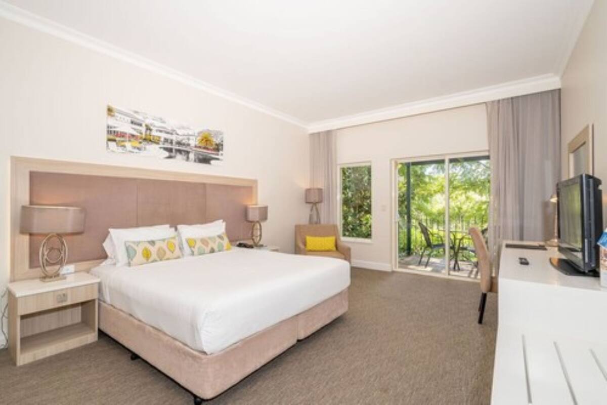 A hotel room with a large bed, two bedside tables, a desk with a chair, a TV, and a large window with a view of greenery.