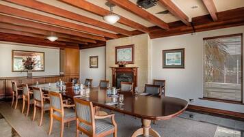 A conference room with a large wooden table, chairs, a fireplace, framed artwork, and a window with blinds.