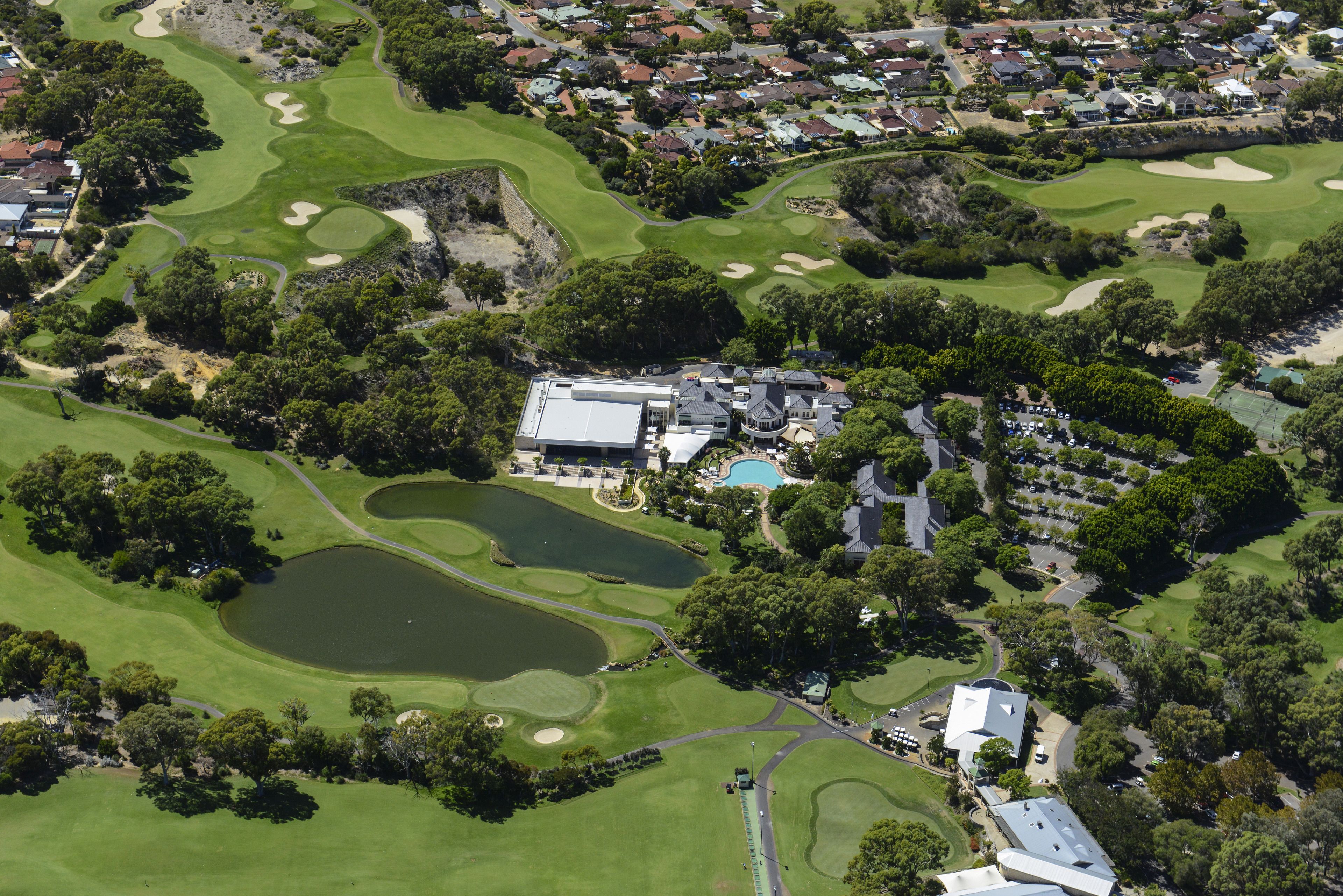 An aerial view of a golf course with a clubhouse, multiple ponds, and residential areas in the background.