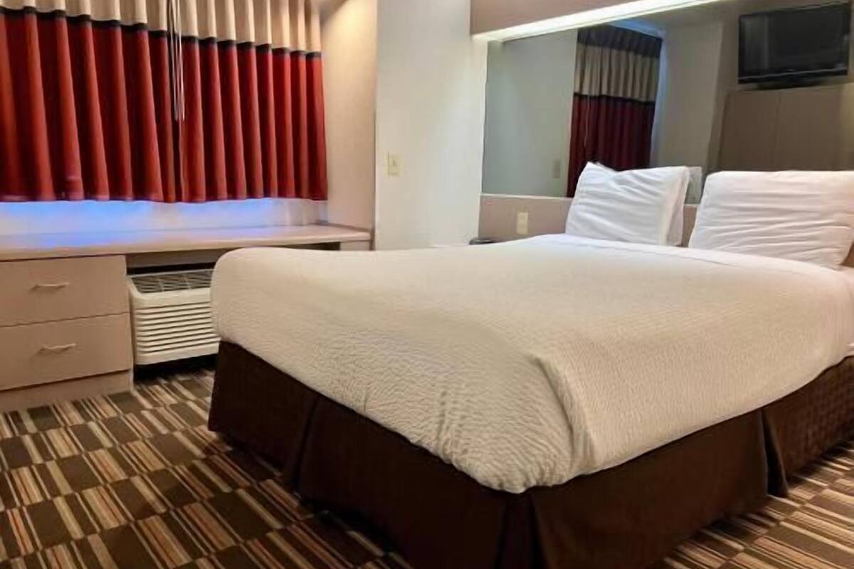 Une chambre d’hôtel avec un lit, un bureau, un miroir et une fenêtre avec des rideaux.