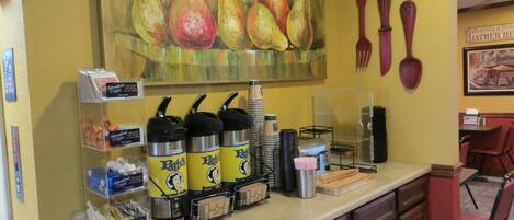 A breakfast bar with coffee dispensers, a painting of pears, and a sign that says "Eat".