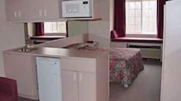 Una cocina compacta con microondas, un refrigerador pequeño y un lavamanos. Hay una ventana con cortinas y una cama con colcha estampada floral.
