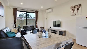 A modern living room with a dining area, a flat-screen TV mounted on the wall, and a large window with curtains. - Comfort Hotel Dandenong (Dandenong)