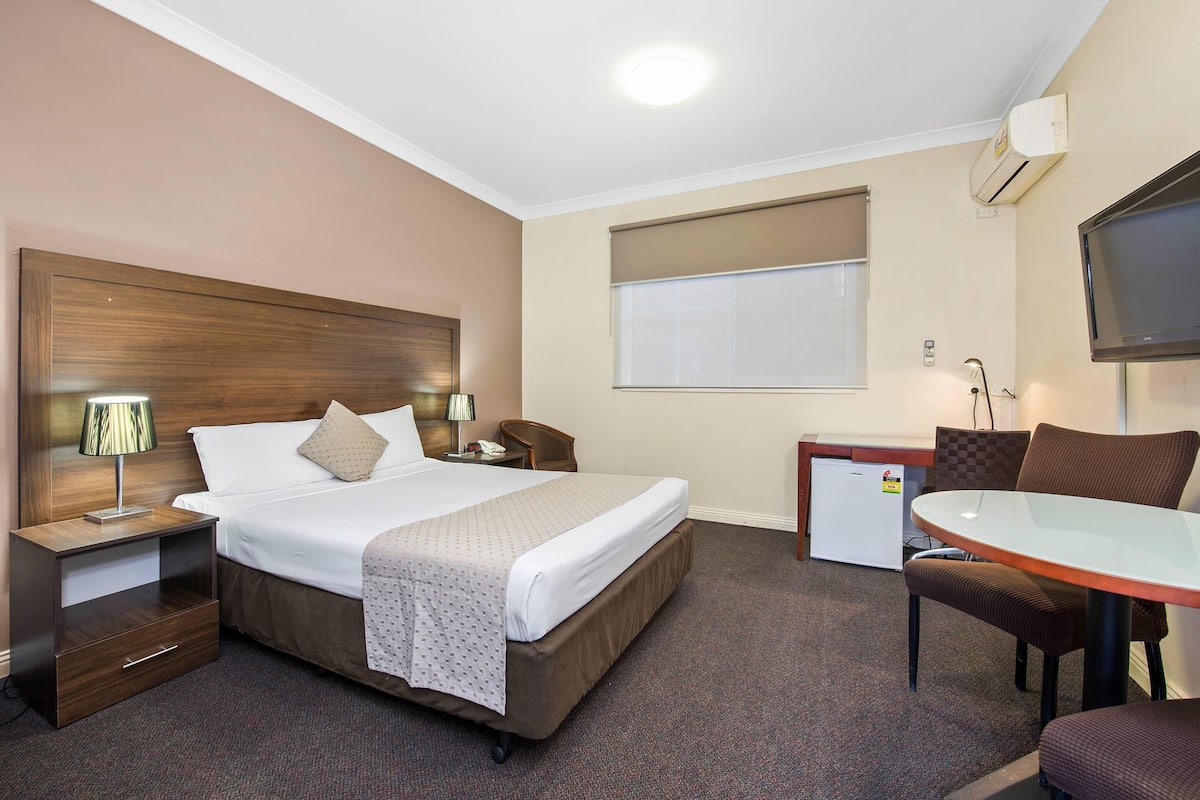 Hotelkamer met een bed, nachtkastjes, een bureau, een stoel, een televisie en een raam met jaloezieën.