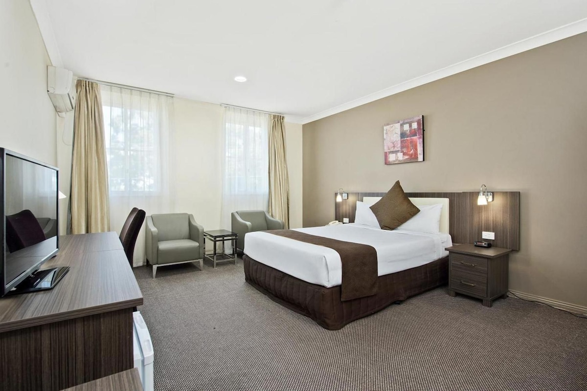 A hotel room with a bed, a desk with a TV, two chairs, a bedside table with a lamp, and a painting on the wall.