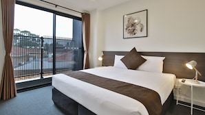 A hotel room with a large bed, a nightstand with a lamp, a framed artwork on the wall, and a view of the outside through a window with curtains. - Comfort Hotel Dandenong (Dandenong)