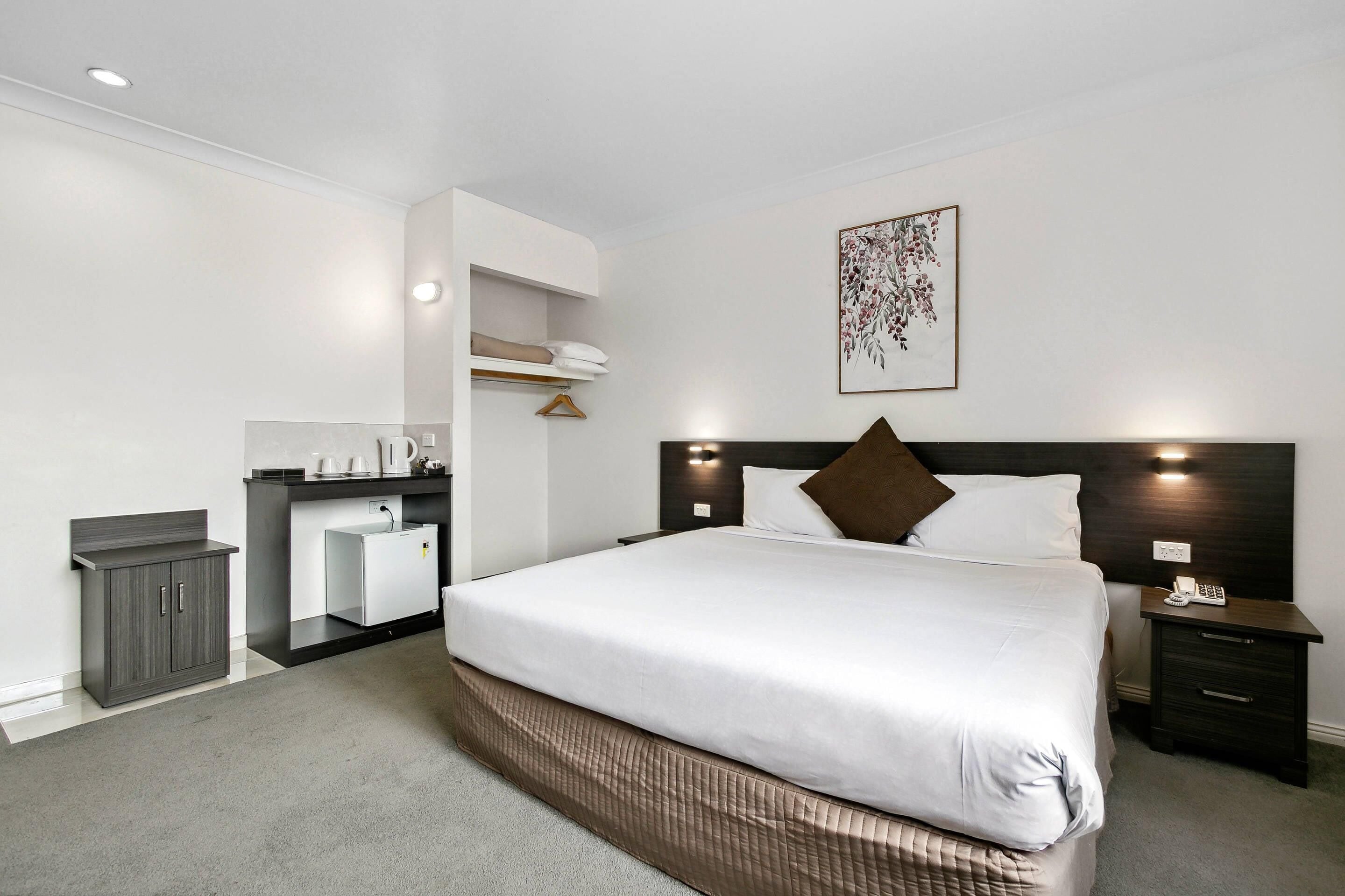A hotel room with a bed, a bedside table, a nightstand, a small desk with a chair, a wardrobe, and a wall-mounted shelf.