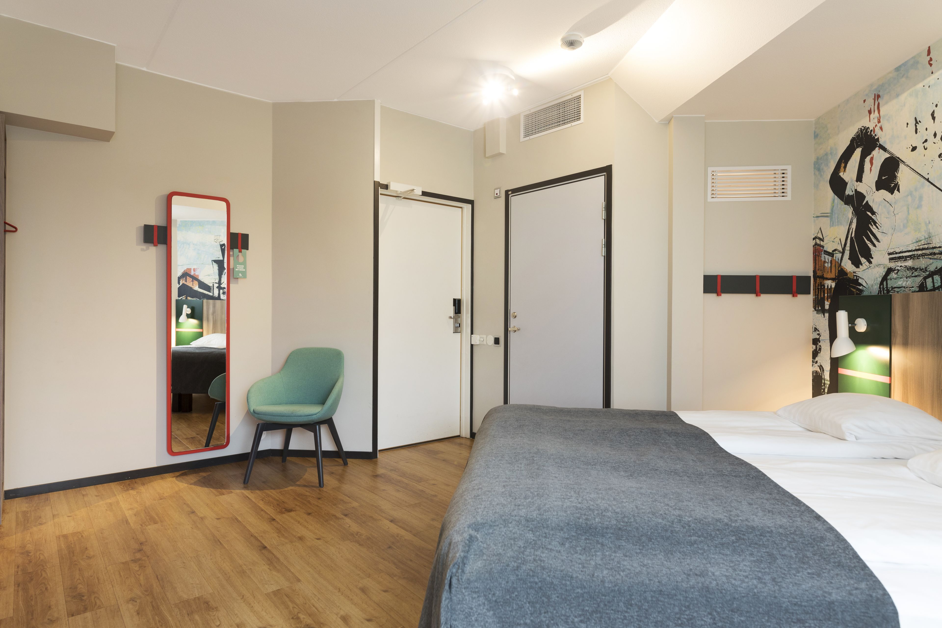 Ett modernt hotellrum med trägolv, en säng med ett grått överkast, en grön stol, en stor spegel och en vägg med en tavla.