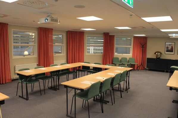 Un salón de conferencias con mesas largas y sillas, un proyector y ventanas con cortinas.