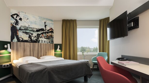 Een hotelkamer met een bed, een bureau met stoel, een televisie en een groot schilderij aan de muur.