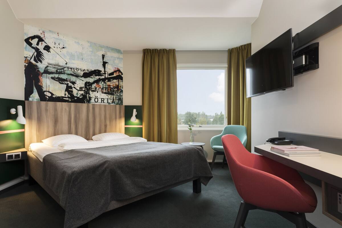 Una habitación de hotel con una cama, un escritorio con una silla, un televisor y un cuadro grande en la pared.
