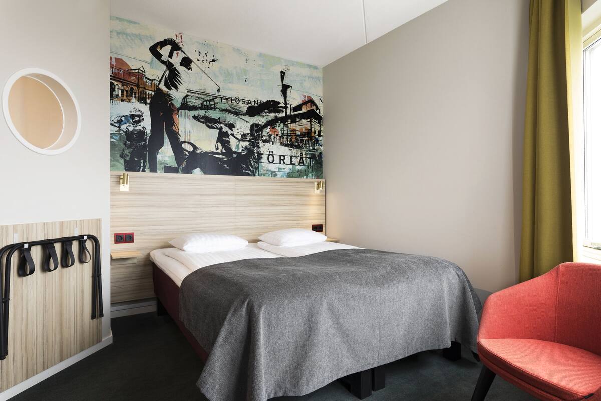 Habitación de hotel con cama, una silla roja, una obra de arte en la pared y un espejo redondo.