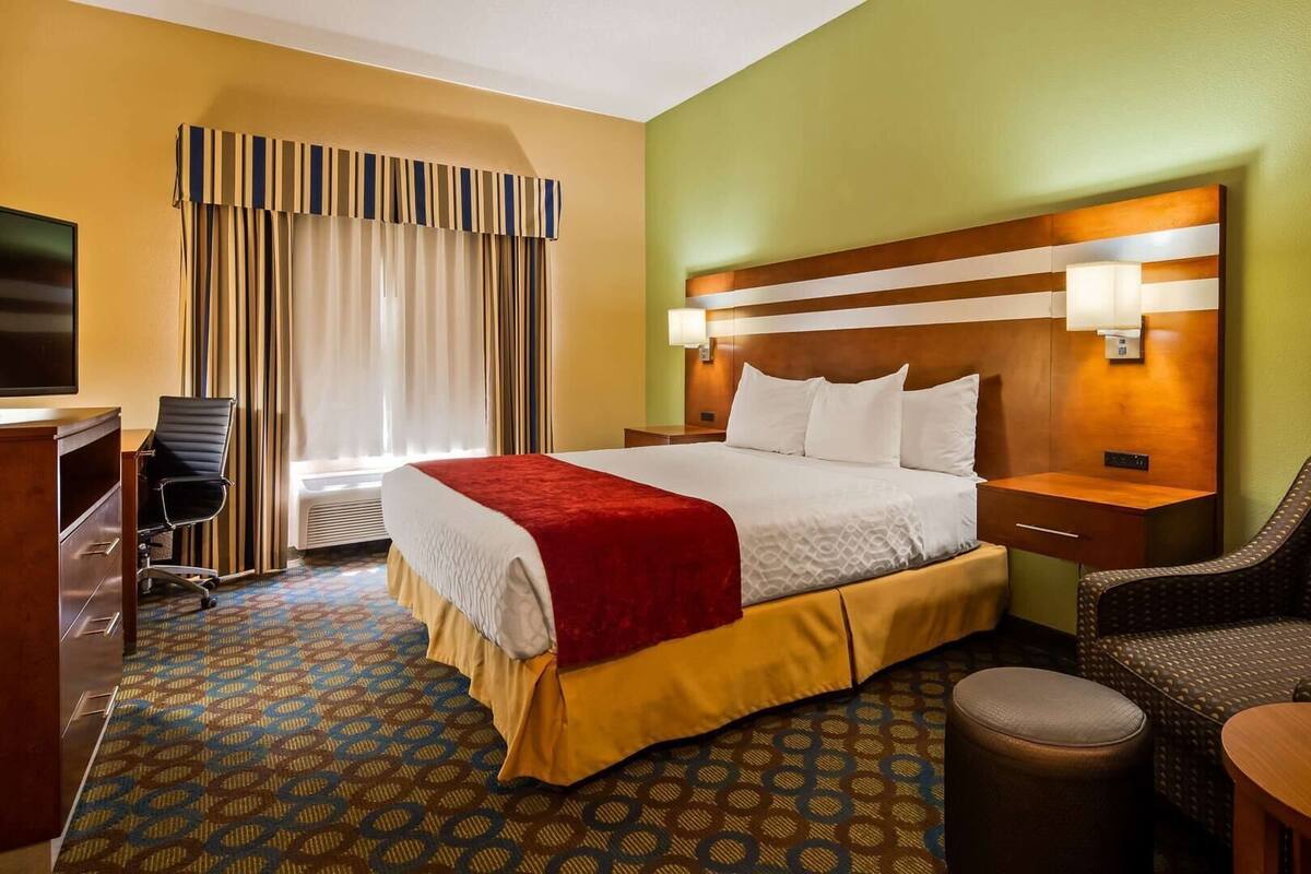 A hotel room with a bed, a desk, a chair, a lamp, and a TV.