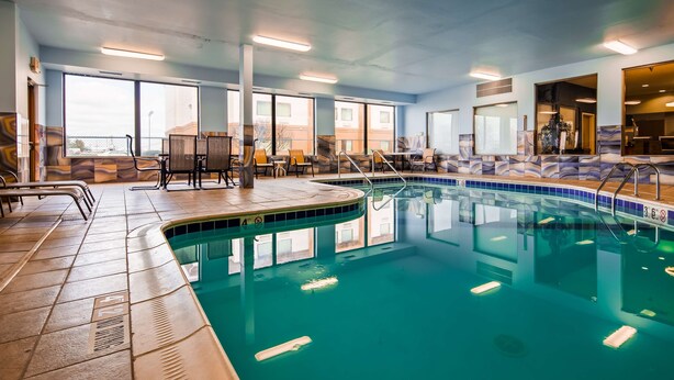 An indoor swimming pool with clear water, surrounded by lounge chairs and tables, and large windows offering views of the outside. - Best Western Troy Hotel (Troy)