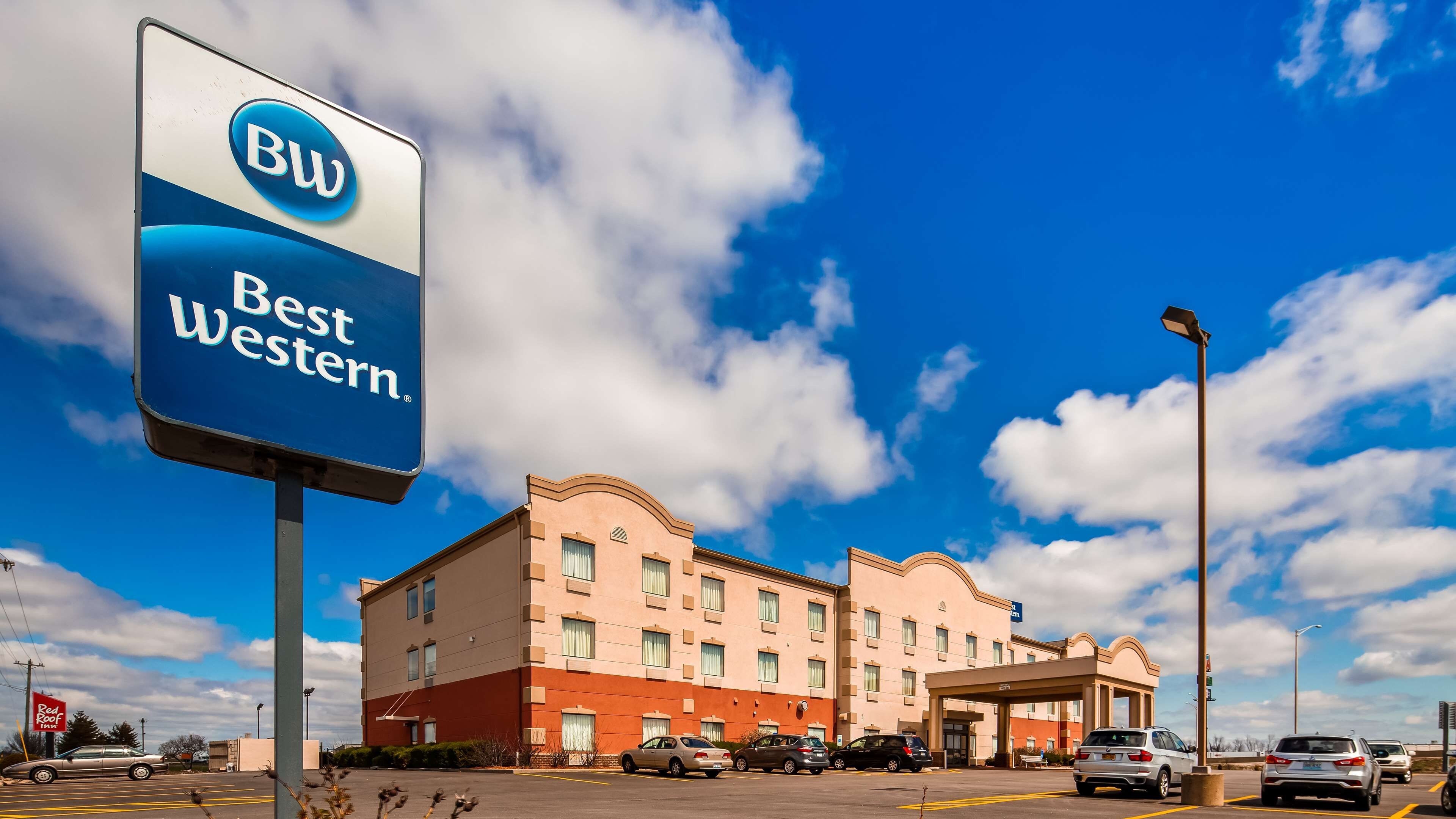 Um hotel Best Western com fachada vermelha e bege, vários espaços de estacionamento e um céu azul límpido.