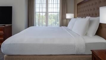 A large bed with a tufted headboard, a flat-screen TV on a wooden dresser, and a window with curtains.