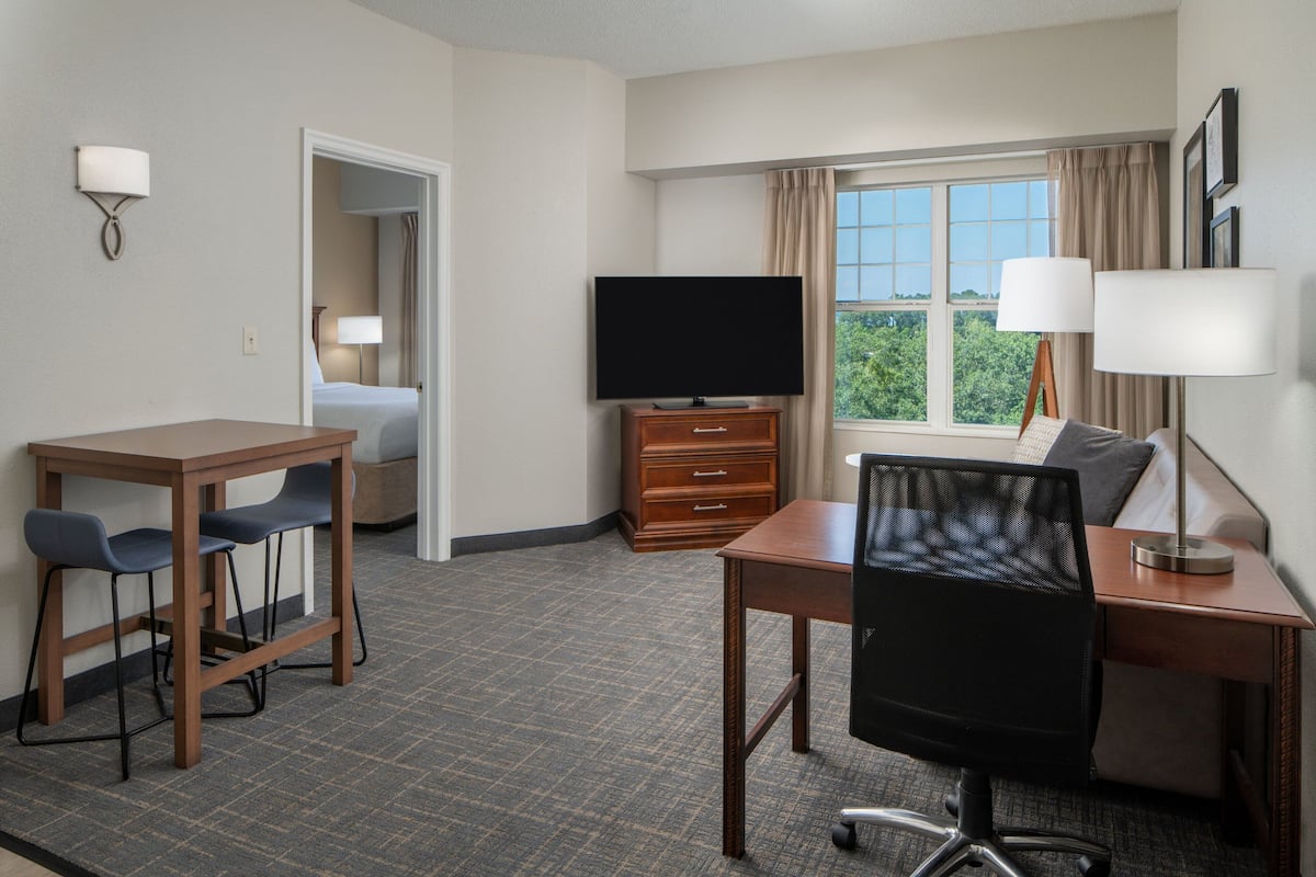 Ein Hotelzimmer mit einem Bett, einem Schreibtisch mit zwei Stühlen, einem Fernseher, einem Schminktisch und einem Fenster mit Vorhängen.