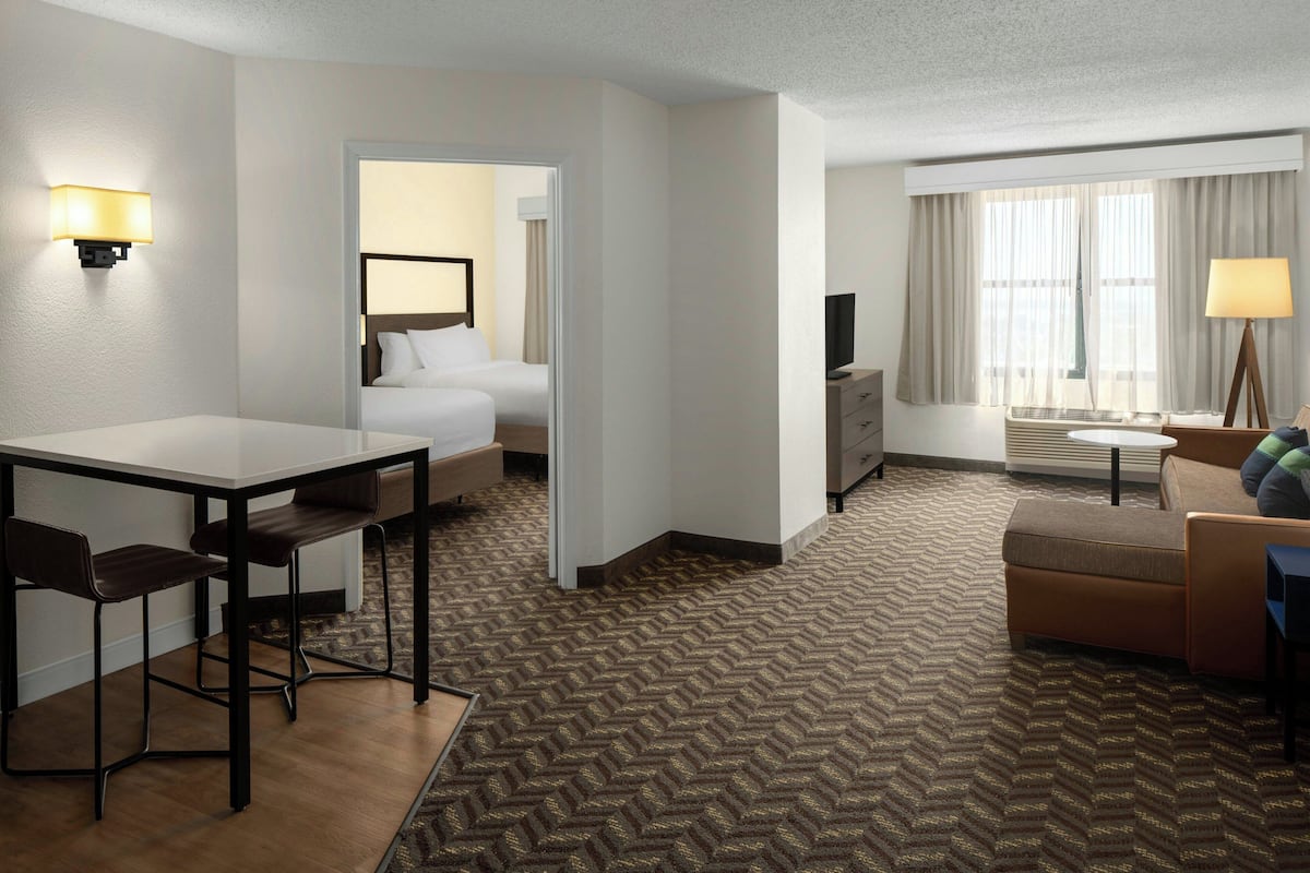 A hotel room with a bed, a desk with two chairs, a sofa, a TV, and a window with curtains.