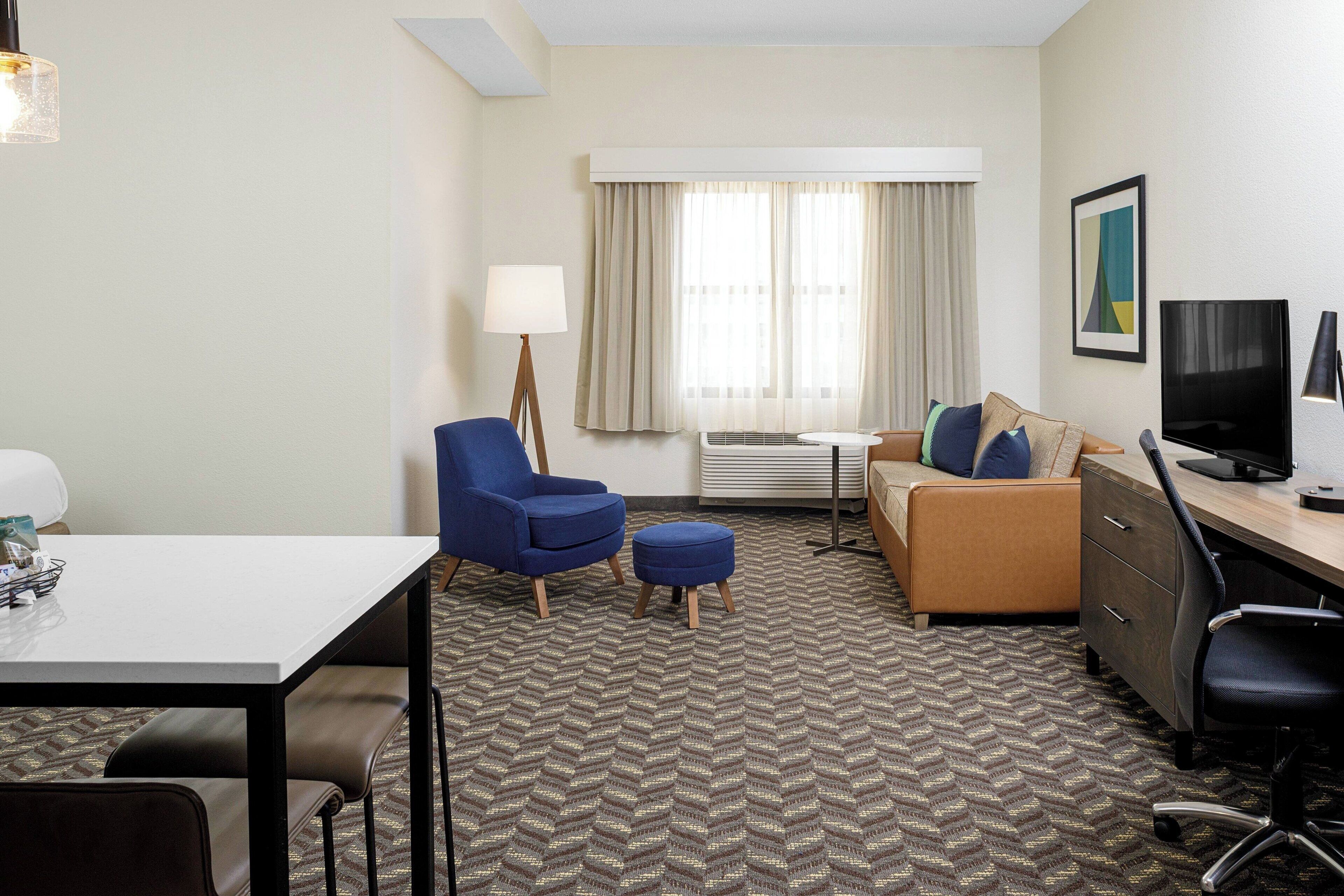 Habitación de hotel con un escritorio blanco, un sillón azul, un sofá marrón, un televisor y una alfombra estampada.