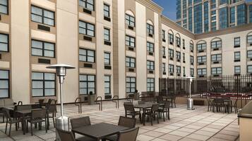 An outdoor patio area with multiple tables and chairs, a large building with arched windows, and a tall skyscraper in the background.