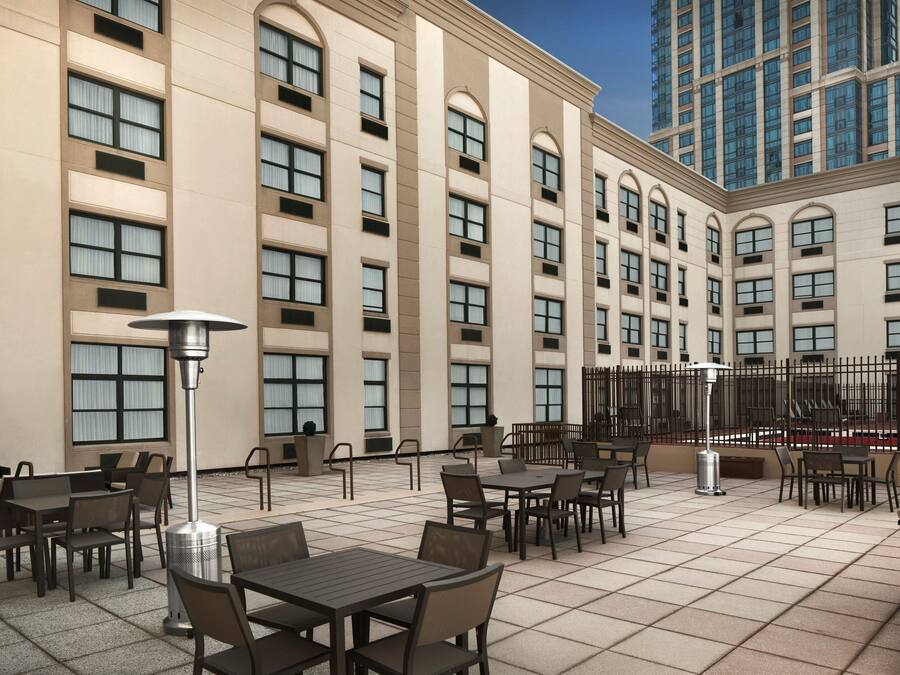 An outdoor patio area with multiple tables and chairs, a large building with arched windows, and a tall skyscraper in the background.